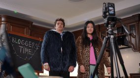 Bottom angle view of cheerful friends gesturing recording video blog indoors on camera. Happy smiling Caucasian boy and Asian girl in fur coats having fun grimacing filming vlog