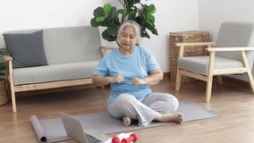Asian elderly woman sitting at home exercising, doing exercises according to online fitness trainers. through a video call on a laptop. Social distancing, maintaining the health of the elderly