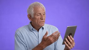 Elderly gray-haired man 70s in blue shirt using tablet computer for telehealth video conference with doctor isolated on solid purple background studio portrait 