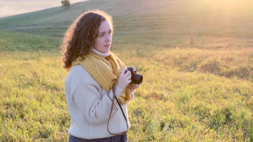 Portrait of curly woman photographer outdoors. Female walk and take pictures with her camera in yellow sunset field.