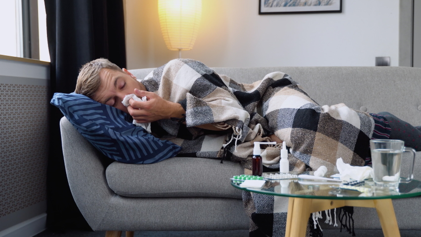 Period of seasonal diseases. Close up of a sick man with flu, fever and headache lying wrapped in a plaid on the couch. A man has runny and common cold. Concept of viral diseases | Shutterstock HD Video #1094826287