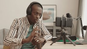 Medium shot of young African American male rap singer wearing earphones sitting at daw at home, rapping while recording it on smartphone camera