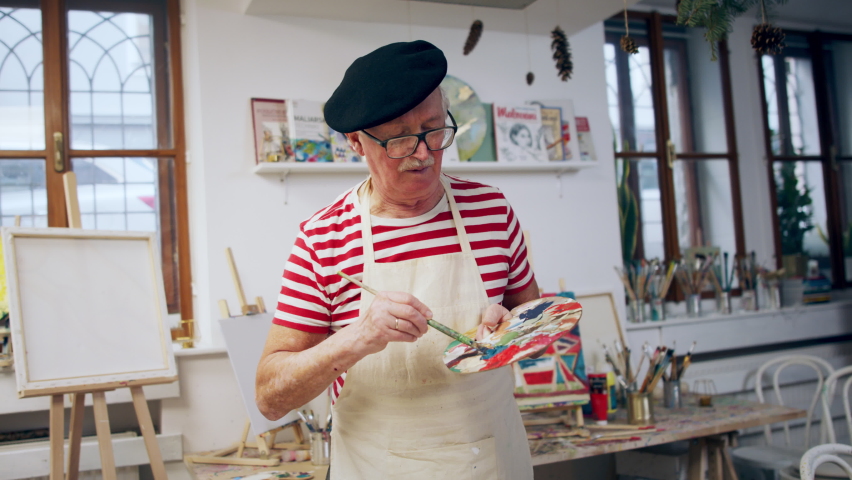 Happy senior man with baret and painting pallete at artist atelier, looking at camera and smiling.