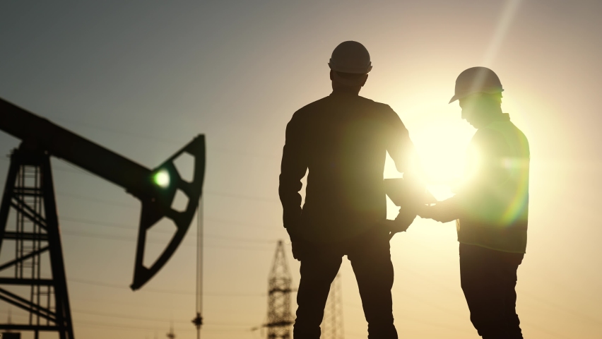Oil production. two silhouette workers work as a team next to an oil pump. business oil production production concept. two engineers of sun the oil and gas industry are discussing a business plan