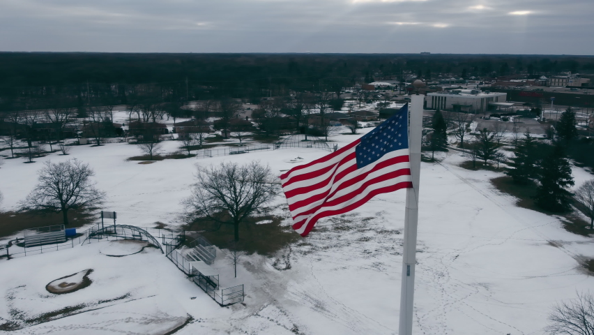 A drone flies around the US Flag in winter in Illinois. The USA flag flies in the center of the village in cloudy weather in winter. | Shutterstock HD Video #1094844441