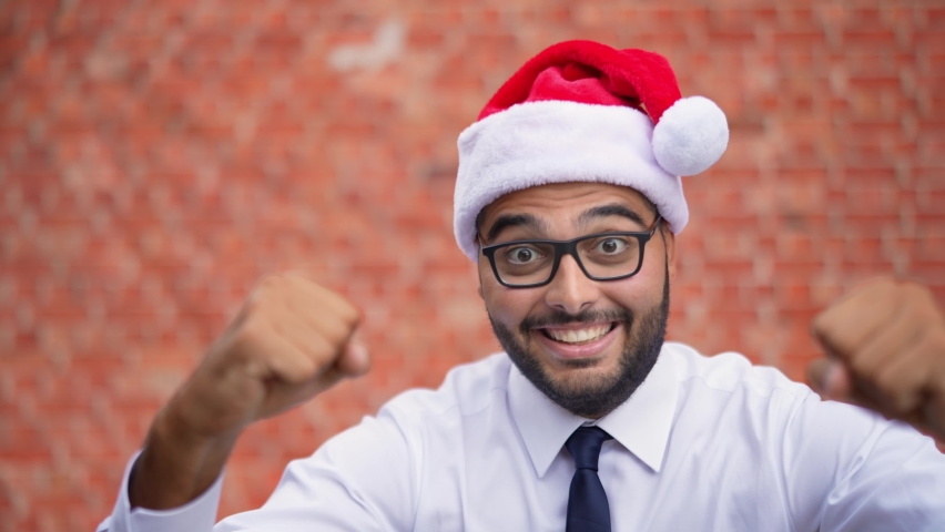 Portrait of a happy young guy in a white shirt, tie, and a Santa Claus hat enjoys success and celebrates victory, gesturing happily. | Shutterstock HD Video #1094849117