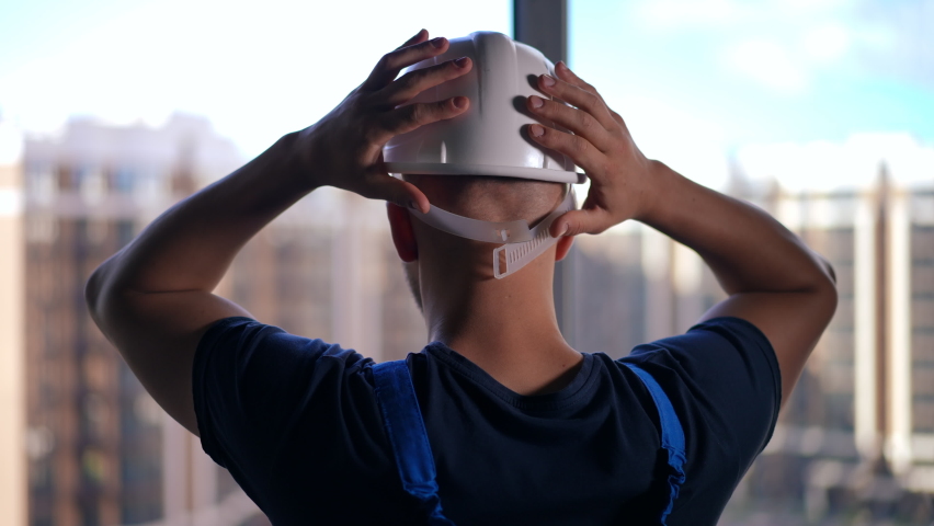 Back view close-up builder putting on white hard hat standing at window looking out at residential neighborhood. Unrecognizable Caucasian young man in uniform indoors in building under construction