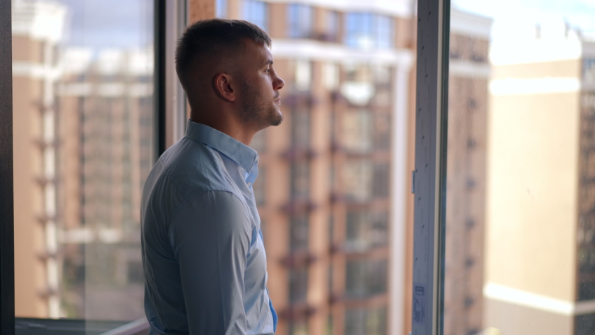 Caucasian young man looking out the window crossing hands smiling standing in new flat. Side view of confident satisfied apartment owner admiring neighborhood in the morning looking around