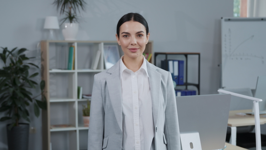 Footage of smiling female CEO of company in elegant gray business suit looking at camera. Open space on office. Portrait of successful young charming feminine woman. Manager, office. Arms folded Royalty-Free Stock Footage #1094853945
