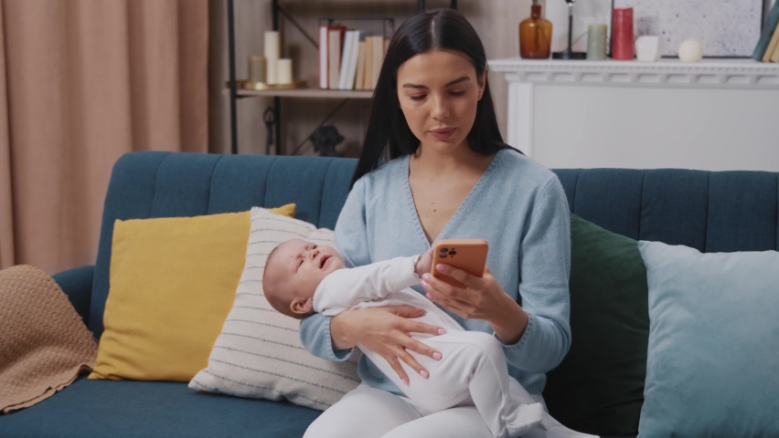 Close up busy mother use phone holding on arms newborn baby crying sitting on sofa in modern apartment. Motherhood. Childhood. Family. Smartphone scrolling. Slow motion Royalty-Free Stock Footage #1094854067