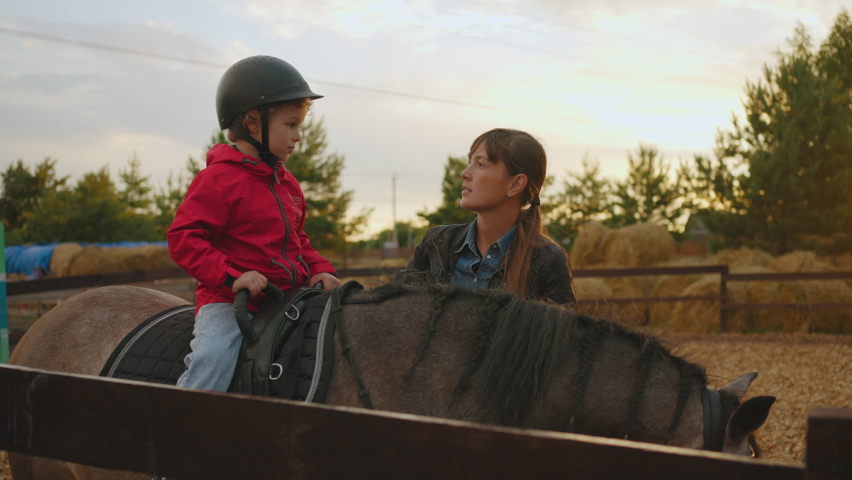 Therapeutic riding instructor woman is talking with little boy on horseback in equestrian club during training | Shutterstock HD Video #1094854313