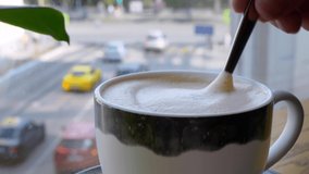 A mug of cappuccino is on the table by the window, which is stirred with milk foam with a spoon, out of focus cars pass by the window. Busy city life when you need a coffee break.