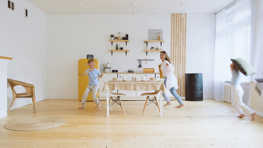 Mother and children have fun running at home around kitchen table fighting on pillow. Leisure of mom, son, daughter, true emotions. Happy merry family, parents play with small kids. | Shutterstock HD Video #1094856955