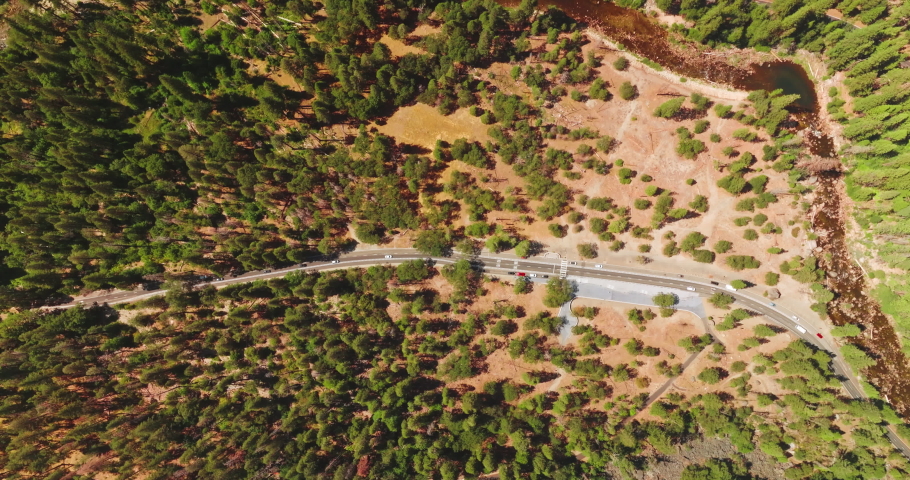 Cars going by the road in Yosemite National Park, California, United States. Pine tree forest growing on the dry rocky landscape from top view.