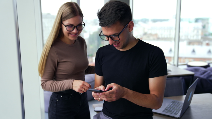 Young diverse employees in office stand at the window looking at phone. Lady telling something to a man typing on the smartphone. Blurred backdrop. | Shutterstock HD Video #1094857911
