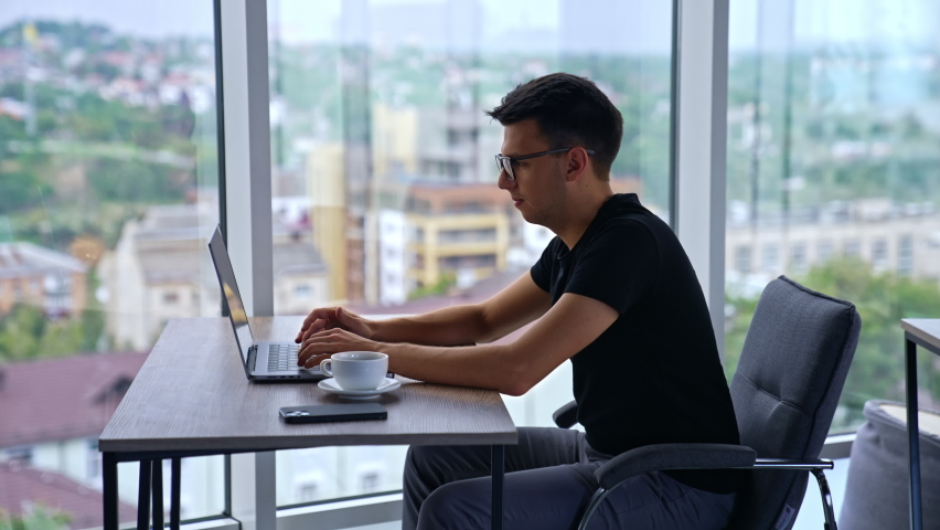 Young adult office employee working at laptop. Dark-haired man focused on work. Blurred cityscape in window at backdrop. | Shutterstock HD Video #1094857925