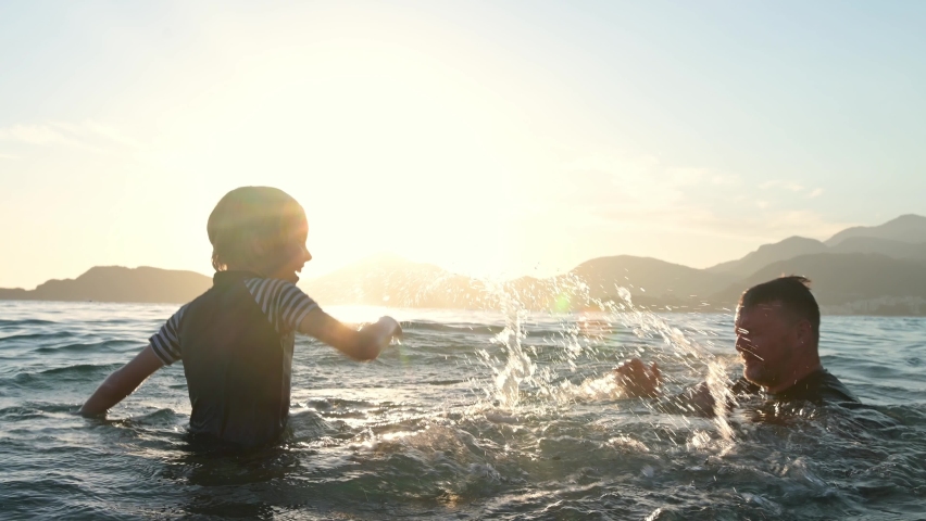 Happy child and his mature father having fun together during swim in the sea at sunset. Joyful boy and his dad splashes with water while swimming in the lake.Active leisure for family during holidays | Shutterstock HD Video #1094858939