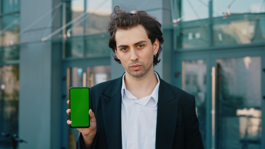 Young businessman showing a smartphone with a green screen while standing near the work place. Caucasian man pointing at a smartphone with a chroma key | Shutterstock HD Video #1094862469
