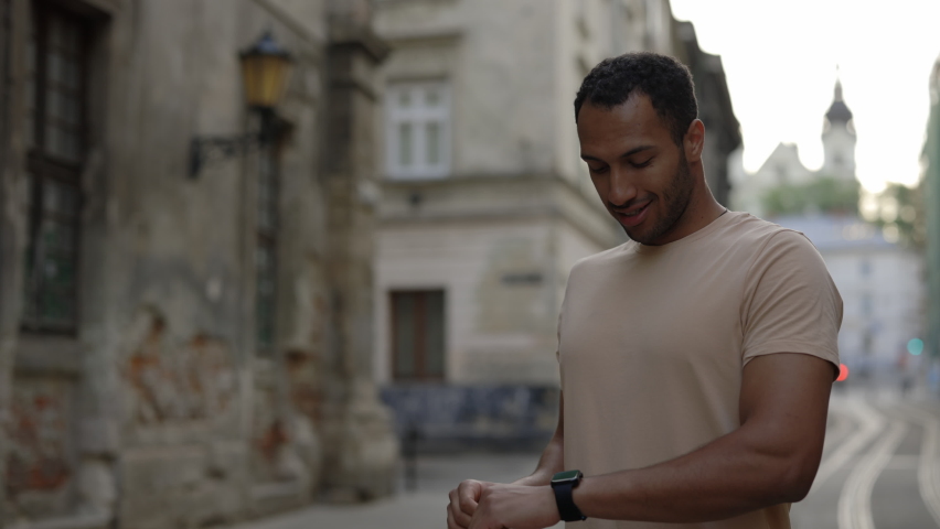 Portrait of active african american man using activity tracker on smartwatch before run workout, standing in the old town street. Sport and healthy lifestyle concept | Shutterstock HD Video #1094878563
