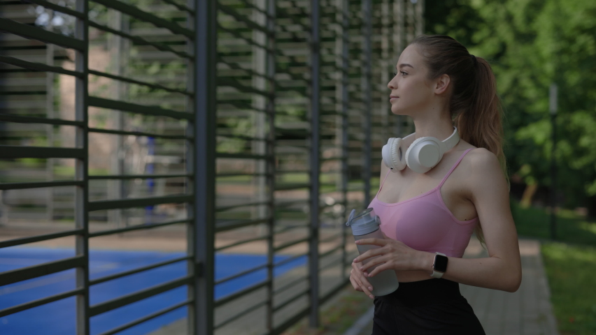 Portrait of the young sport woman wearing a headphones on her neck holding the sport bottle and wearing the smart watch looking at the camera. Sport and healthy lifestyle concept | Shutterstock HD Video #1094878569