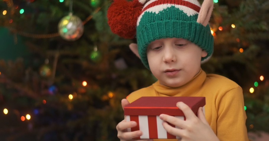 Boy in Christmas elf hat opens Xmas presents. Child with joy opens gift box sitting under the Christmas tree