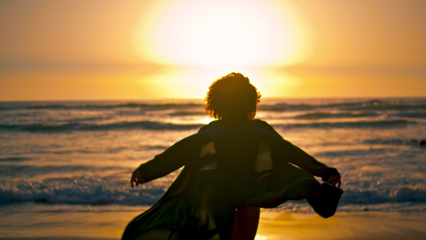 Back view of african american woman walking at sunrise on ocean waves. Unknown attractive girl in black swimsuit raising hands enjoying soft sunlight. Slim young lady relaxing on beautiful nature,
