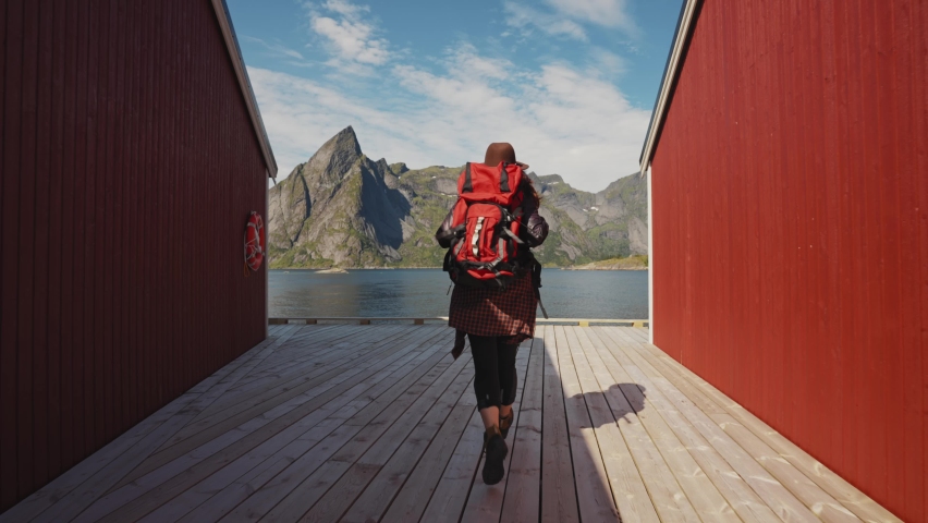 red head girl runs on the pier towards the sea next to the traditional red wooden rorbu fisherman