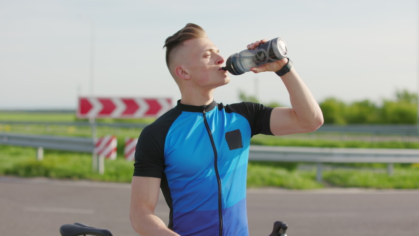 Athlete man drinking water from bottle after training on professional bicycle. Healthy active man resting on side of road and looking at amazing landscape. | Shutterstock HD Video #1094898225