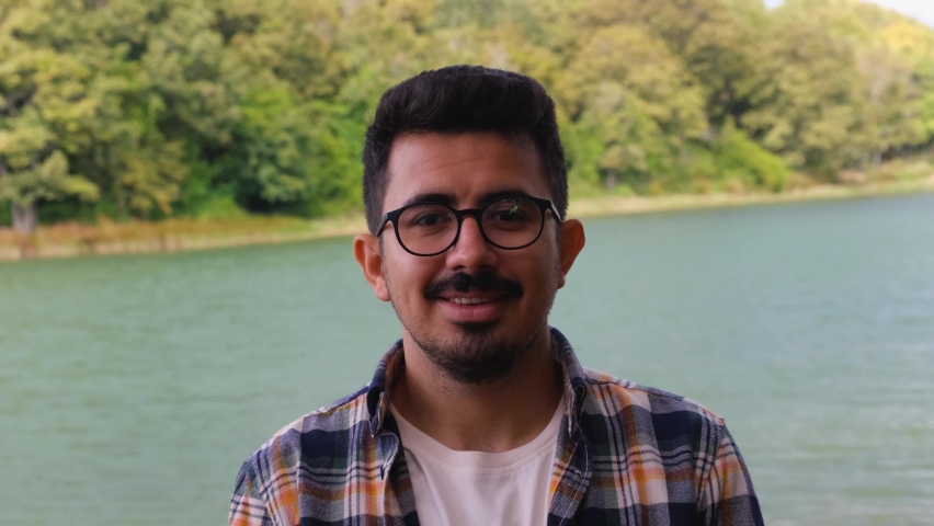 Smiling young man with moustache and beard, man with glasses laugh front of lake, Turkish bearded person, 4k 60 fps video of portrait with nature landscape in background, brunette male with a shirt | Shutterstock HD Video #1094902873