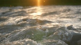 Afternoon sun shines on rapid river waves, drops of water glowing in air, closeup slow motion video