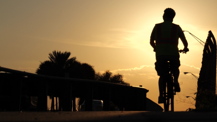 Male cyclist in silhouette rides toward the sunset up and down the sidewalk along the Florida Turnpike overpass at Sample Road in Pompano Beach | Shutterstock HD Video #1094908429