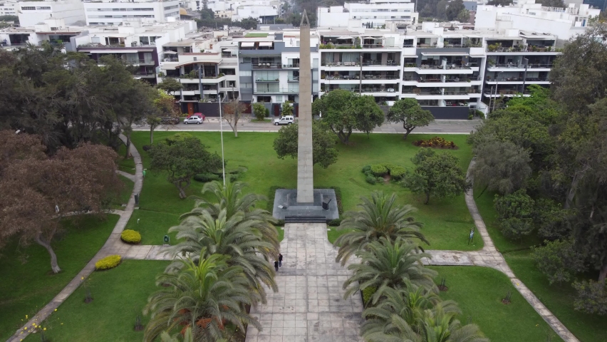 Drone video of an obelisk in a park. Many trees and green grass around. Drone flies high above and towards the peak of the monument. Recorded in Lima, Peru. In San borja district. In "Parque Aramburu"