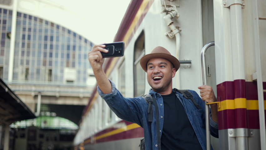 Freedom traveler young asian man hanging handrail on train and taking photo by smartphone. Happy tourist travel by train on vacation time holiday weekend trip. Backpacker arrival at platform railway.