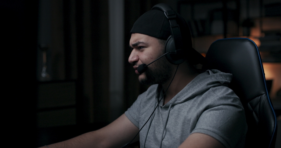 Guy with beard in gray t-shirt and black cap is playing online game. Player emotionally communicates with game partner, smiles. Man is sitting on chair, wearing headphones and holding computer mouse.