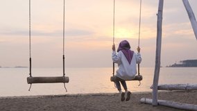Girl in an oversized sweatshirt swings on a wooden swing at sunset near the sea. Back view

