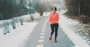 Running woman, headphone girl runner on the snow in winter sunny day. Female in pink sportswear jacket fitness training outdoors. Sport concept, leisure and freedom. 4K slow motion video