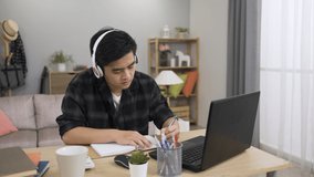 portrait of a concentrated Asian man with earphones on head is repeating after the teacher and writing notes while taking online classes on the computer at home