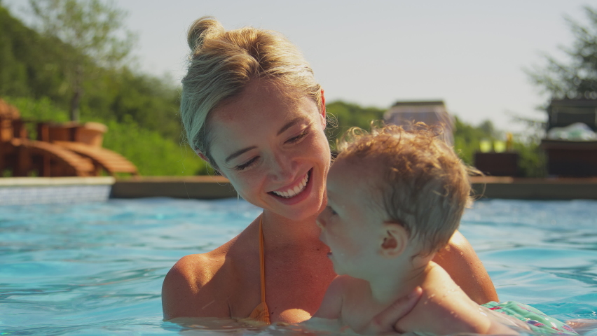 Smiling mother with baby son on family summer holiday relaxing and splashing in swimming pool - shot in slow motion