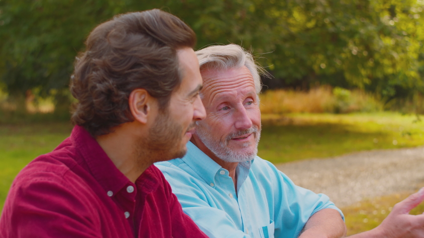 Casually dressed smiling mature man with mixed race adult son leaning on wooden fence on walk in summer countryside talking together - shot in slow motion