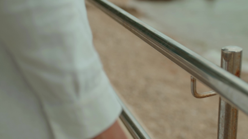 The young girl in a long white dress walks near the sea and rolls her hand along the metal railing