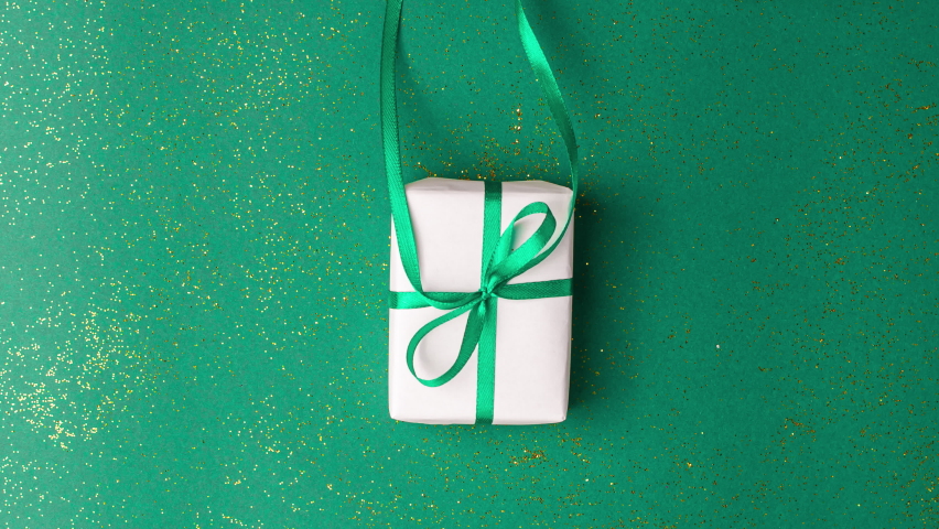 Human hands in white sweater take a white paper gift with a emerald green satin ribbon bow on green background with gold colored glitter. Stop motion animation Christmas Holidays concept flat lay. | Shutterstock HD Video #1094936781