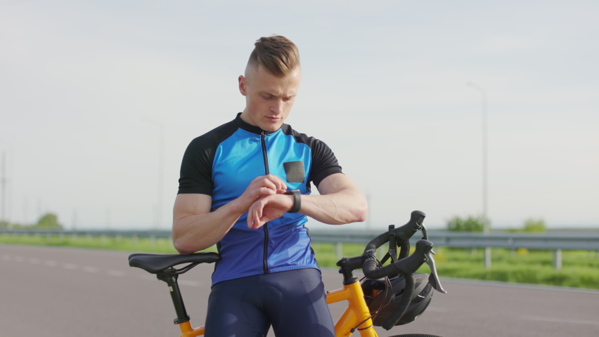 Caucasian sportsman in uniform using smartwatch app for fitness tracking after workout training outdoors. Young handsome cyclist standing near bicycle at highway. | Shutterstock HD Video #1094937119