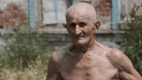 Shirtless elderly man looks in camera on blurred background. Skinny pensioner with bare torso stands in yard against shabby brick house in village closeup - Powered by Shutterstock - Get 15% off with code: PIKWIZARD15