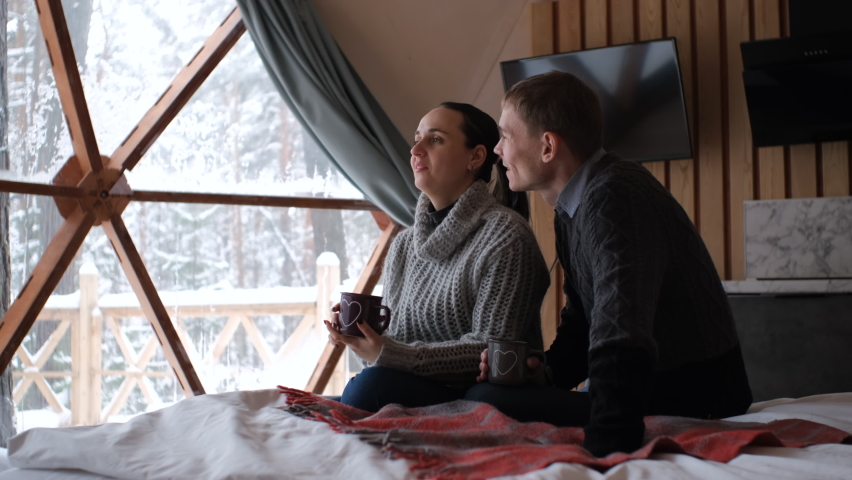 Young couple drinking tea or hot chocolate while sitting in bed in dome camping during winter holidays. Comfort and tranquility glamping vacation concept | Shutterstock HD Video #1094944317