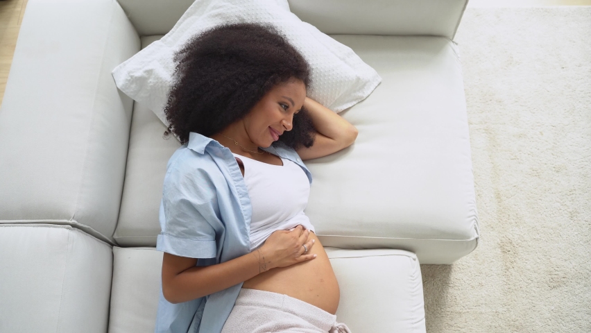 Pregnant black beautiful woman lying on sofa at home, touching belly, dreaming about future. Relax and healthy concept of conscious pregnancy. | Shutterstock HD Video #1094953099