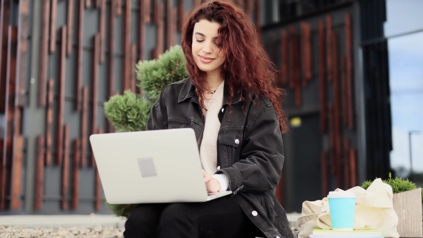 Portrait of attractive curly woman using laptop while sitting on a bench near the modern city buildings. Study, learning, remote work, freelance.  | Shutterstock HD Video #1094954717