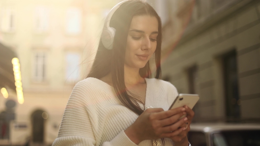 Charming woman with headphones using social media application on smartphone, texting messages, receive news in the city street. | Shutterstock HD Video #1094954719