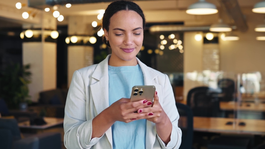 Positive hispanic or brazilian business lady, top manager of a company, standing in her modern creative office, uses her smart phone, messaging in social media, answers email, typing, smiling | Shutterstock HD Video #1094955163