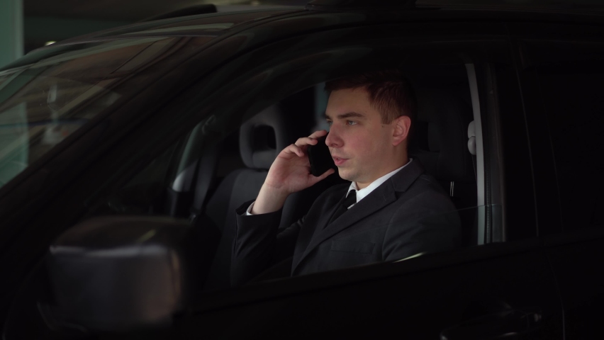 A young businessman sits in a car in the parking lot and talks on the phone. A respectable man communicates via cellular communication. A man in a suit is driving a car. | Shutterstock HD Video #1094955287
