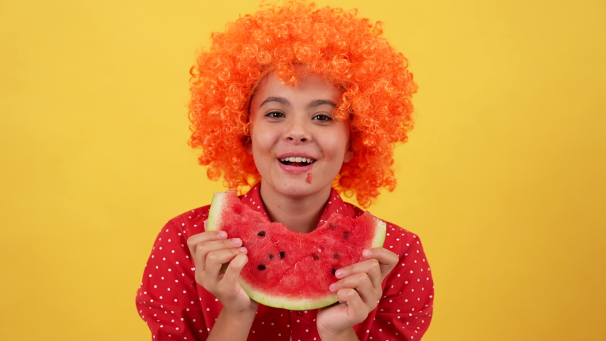 Amazed happy kid in orange hair wig eating slice of water-melon fruit on yellow background, vitamins | Shutterstock HD Video #1094956167